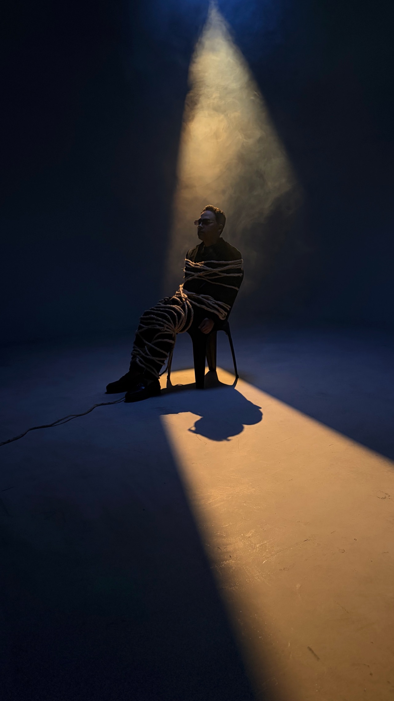 Artista sentado y envuelto en cuerdas bajo un haz de luz cálida en el estudio.
