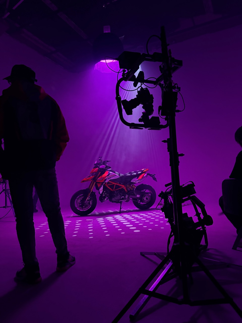 Motocicleta preparada en un set violeta entre cámara, luces y miembros del equipo.